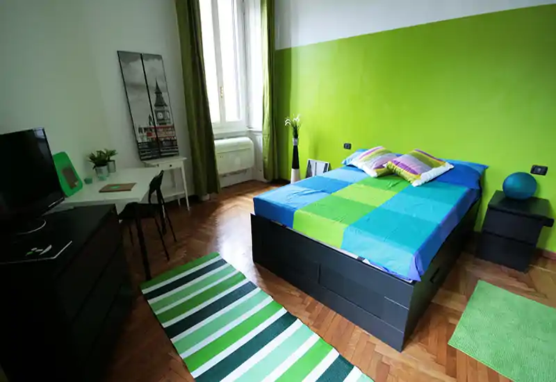 Bright, well-staged bedroom with a double bed dressed in blue and green linens, parquet floor and coordinated rugs; natural light from a window brightens the room.