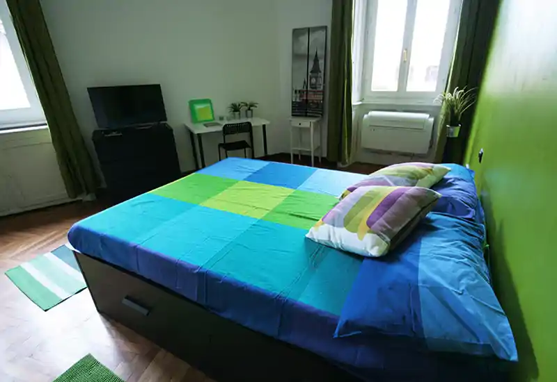 Bright, modern bedroom with a large double bed dressed in colorful linens, two pillows, wooden parquet flooring and a small desk by the window — well staged and airy.