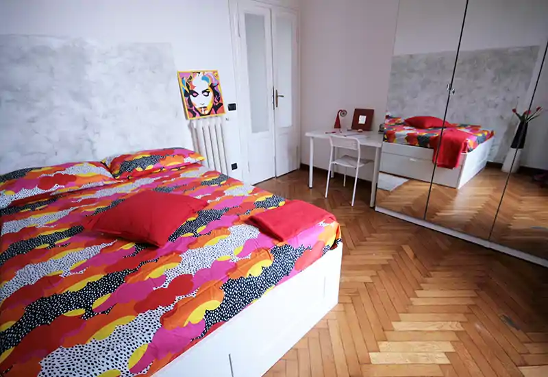 Bright, colorful bedroom with a large double bed featuring vibrant patterned bedding, a mirrored wardrobe that expands the sense of space, and a small desk area — ideal for comfortable stays.