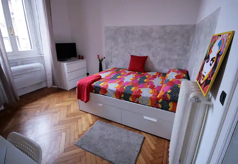 Bright, compact bedroom with a colorful double bed, white storage drawers underneath, parquet flooring and natural light from a large window. The room includes a small dresser with TV and decorative accents for a cozy, stylish stay.