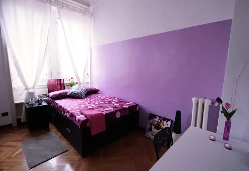 Bright, stylish bedroom with a double bed dressed in purple patterned linens, bedside table, and a desk area — natural light from large windows and coordinated decor create a welcoming atmosphere.