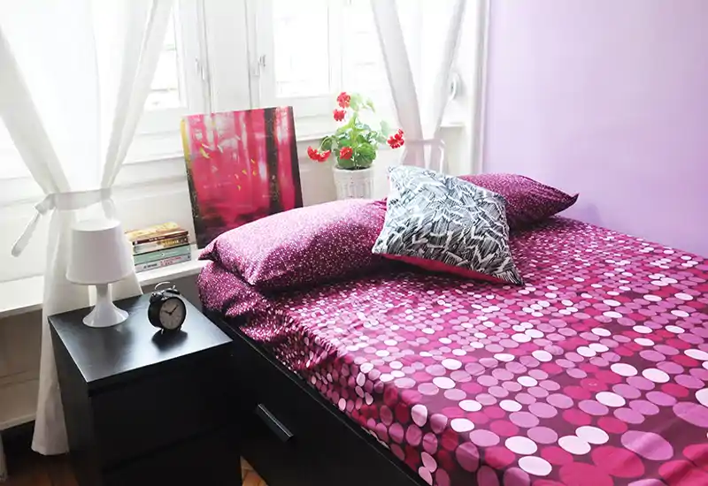 Bright, stylish bedroom with a patterned magenta duvet, decorative pillows and a bedside table with lamp and alarm clock; natural light from large windows creates an inviting atmosphere.