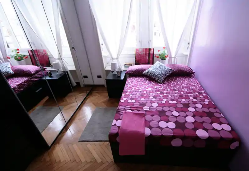 Bright, neatly made bedroom with a patterned pink duvet, bedside tables and mirrored wardrobe that adds depth.
