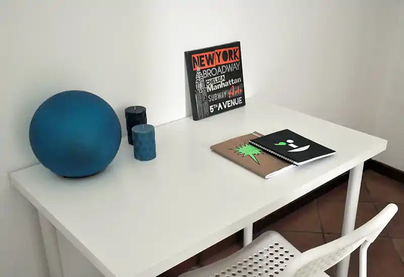 Compact living/work corner with a clean white desk, chair and simple decorative items (blue spherical lamp, candles and a small artwork), suited for a small home office or reading space.