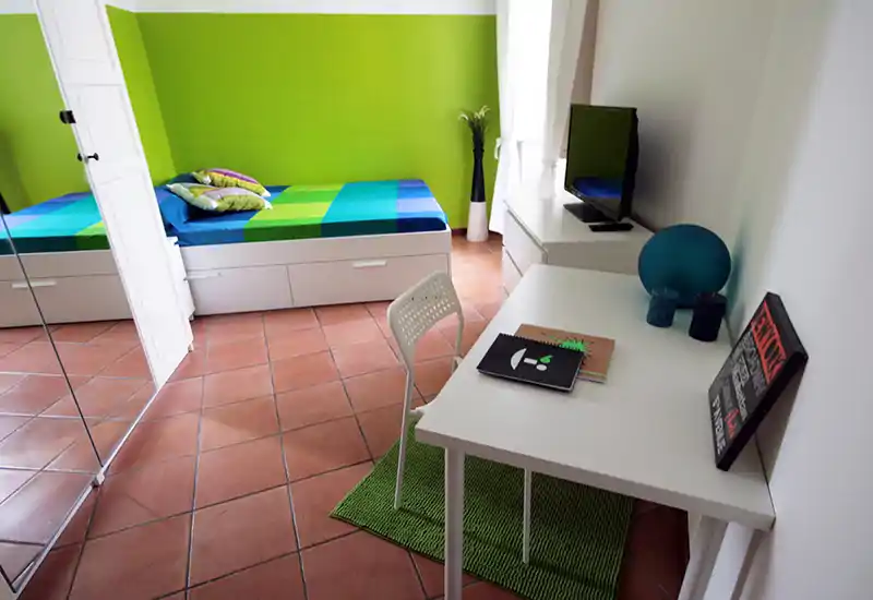 Bright, modern single bedroom with a colorful bed, white furniture and a small work desk by the window — fresh green accent wall and terracotta floor tiles create a lively look.