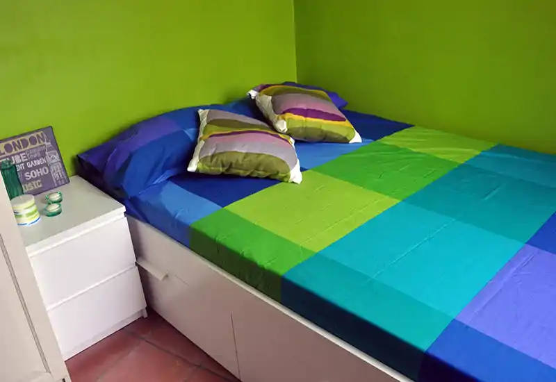 Bright, colorful bedroom with a large bed dressed in vibrant green and blue linens and two decorative pillows. Clean, modern bedside cabinet and tiled floor visible — a compact, inviting sleeping space.