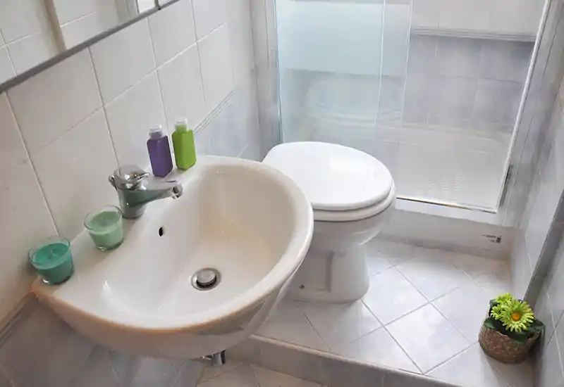 Compact, well-lit bathroom with a wall-mounted sink, toilet and a glass shower enclosure; fresh decorative touches include candles and a small plant.