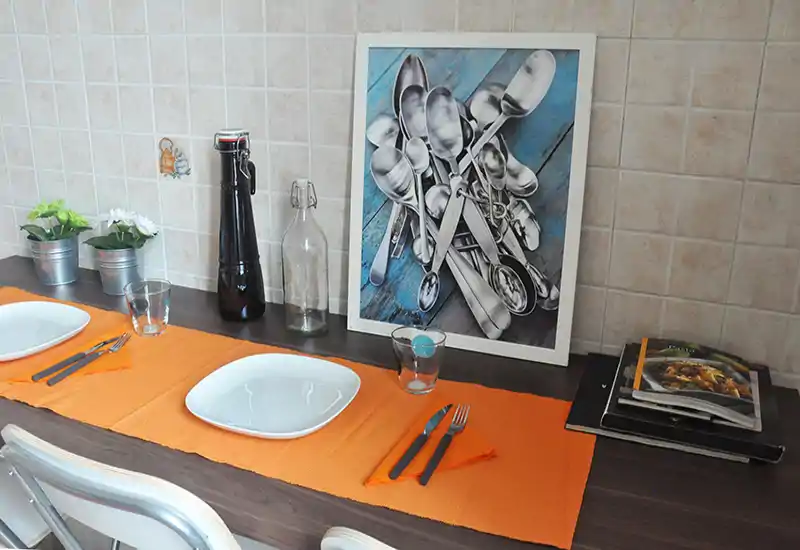 A styled kitchen counter set for two: orange placemats, white square plates, cutlery and glasses on a dark wood breakfast bar with decorative plants and framed artwork.
