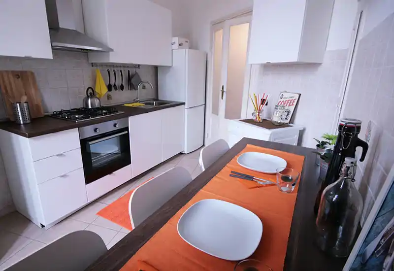 Bright and tidy kitchen with modern white cabinets, gas stove and a wood-toned counter that doubles as a casual dining table set for two. Decorative orange placemats and small plants add a welcoming touch.
