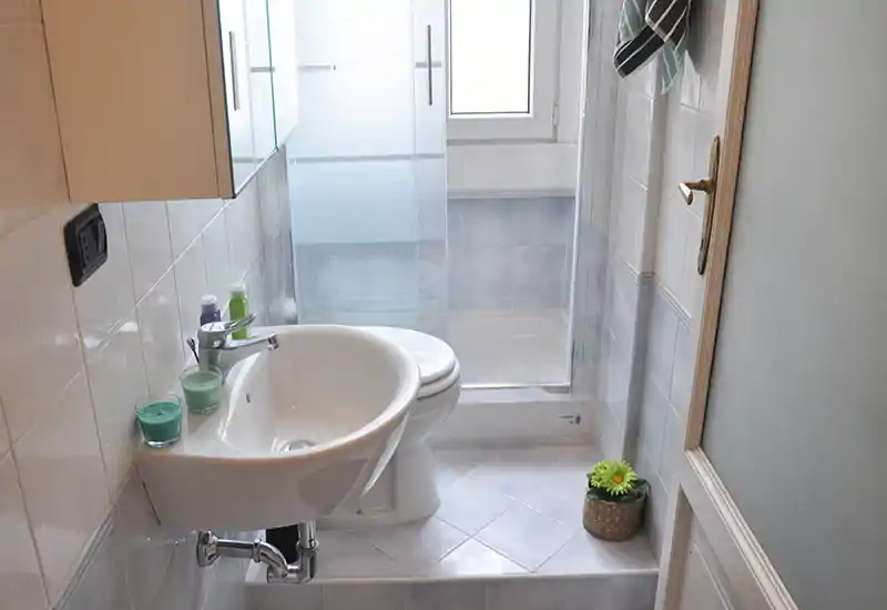 Compact, bright bathroom with a pedestal sink, toilet and a shower cubicle. Neutral tiles and natural light from the window create a clean, functional space.