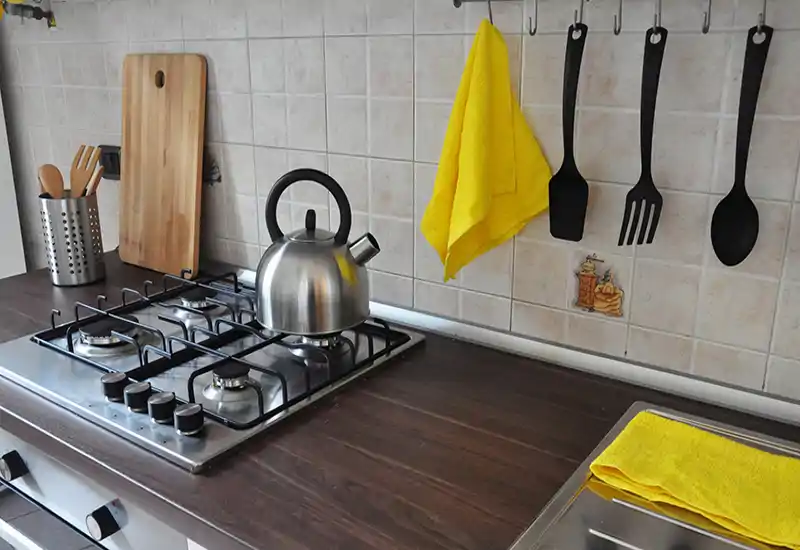 Bright, tidy kitchen counter with a gas hob, stainless-steel kettle and hanging utensils — warm wood countertop and yellow textile accents create a welcoming prep area.