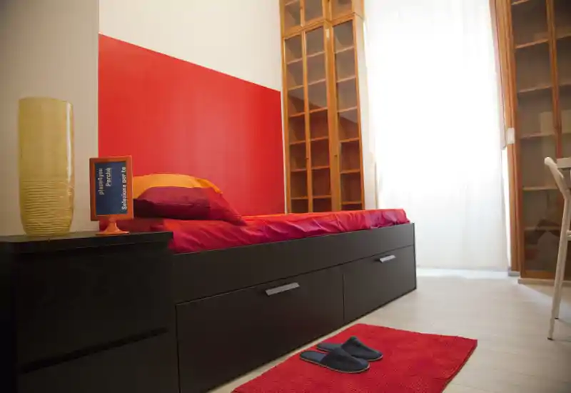 Bright, modern single bedroom with a low black bed frame, vibrant red bedding and a matching rug, lit by natural light from a large window with sheer curtains. Wooden shelving and a small bedside table complete the cozy, contemporary setup.