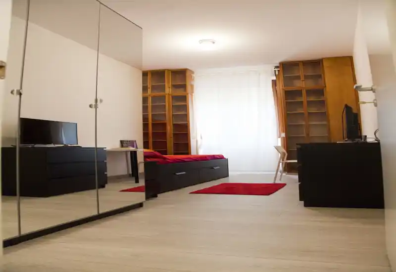 Bright, modern bedroom with a single low bed, red accents and large mirrored wardrobe reflecting the space; wooden shelving and a desk create a functional, cozy layout.