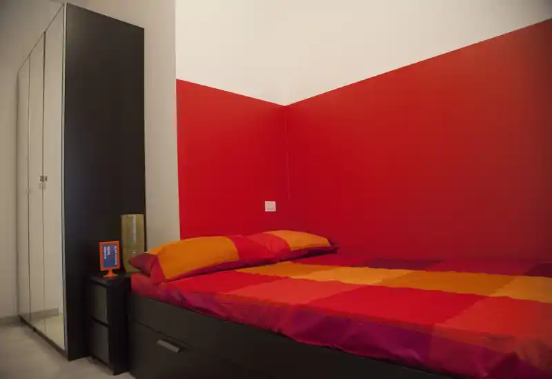 Bright, modern bedroom with a colorful red-and-orange bed and a mirrored wardrobe; compact and well-styled for a single or guest room.