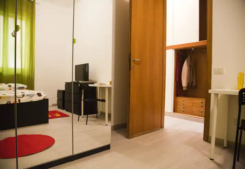 Camera da letto luminosa e ben allestita con letto, armadio con ante a specchio, scrivania e porta in legno che conduce a un'area guardaroba — tessili accoglienti e luce naturale creano un'atmosfera invitante.