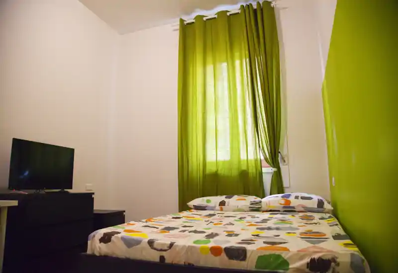 Camera da letto luminosa e ordinata con un letto matrimoniale con copripiumino a fantasia, parete verde e tende abbinate che filtrano la luce naturale. Un comò con TV è di fronte al letto, creando uno spazio compatto e accogliente, ideale per soggiorni brevi.