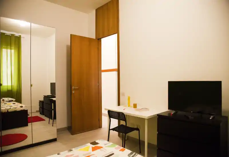 Camera da letto ben illuminata con letto decorato in primo piano, una scrivania bianca con sedia, armadio con ante a specchio e televisore su comò — spazio per dormire semplice, ordinato e funzionale.