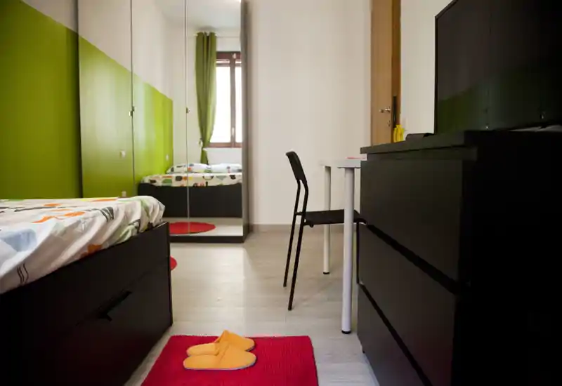 Camera da letto moderna e luminosa con letto singolo, armadio con ante a specchio, scrivania e comò; parete verde e luce naturale la rendono accogliente.