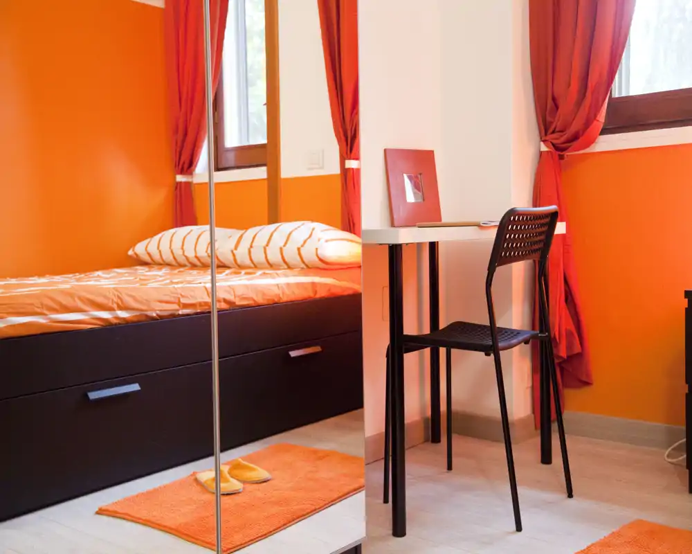 Camera da letto luminosa con tema arancione: letto singolo ben rifatto, tappeto e tende coordinati e una piccola scrivania vicino alla finestra. L'ampio specchio e la luce naturale rendono lo spazio arioso e accogliente.
