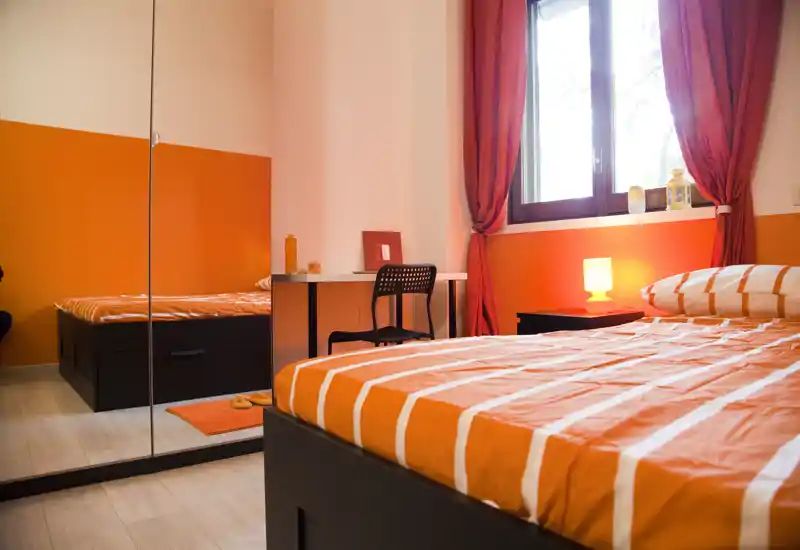 Camera da letto luminosa e ben arredata con un ampio letto a righe arancioni in primo piano, lampada sul comodino e una scrivania vicino alla finestra. L'armadio con specchio e le tende calde aggiungono profondità e un'atmosfera accogliente — perfetta per annunci.