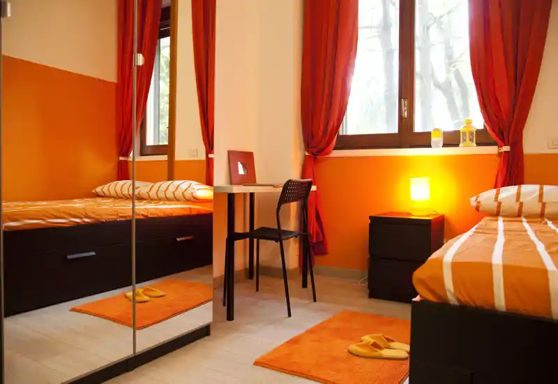 Camera da letto luminosa e accogliente con accenti arancioni — letto singolo con copriletto a righe, tappeti coordinati e tende calde; lampada da comodino e una piccola scrivania accanto alla finestra.