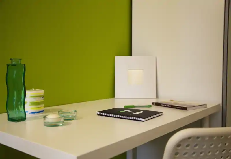 Minimalist desk corner with decorative candles, a green glass vase and a notebook on a white table against a green accent wall — bright, tidy and suited for a workspace or living area.