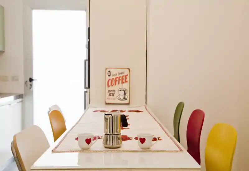 Bright, colorful dining area with a long table set with a runner, coffee pot and heart-motif mugs; playful chairs in yellow, red, green and orange create a cheerful breakfast corner.