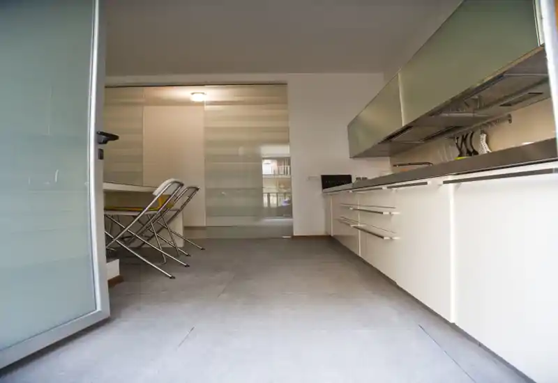 Modern galley kitchen with clean white cabinets, stainless countertop and stools tucked at a small dining nook; bright, minimalist and well organized.