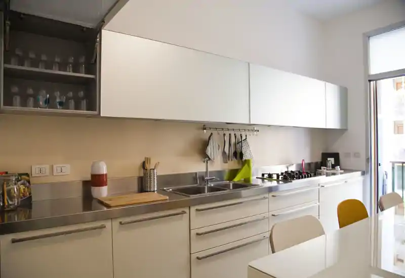 Modern, bright kitchen with stainless steel countertops, sink, gas hob and overhead cabinets. Clean lines and a small dining table create a functional, stylish cooking space.