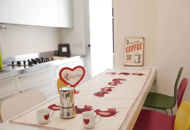 Bright, modern kitchen area with a decorated table runner, espresso maker and two heart-printed mugs; colorful chairs add a playful touch.