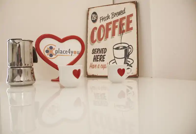 Close-up of a bright kitchen countertop styled as a coffee corner: two small heart-decorated cups, a stovetop moka and a vintage 'Coffee' sign, with warm decorative accents.