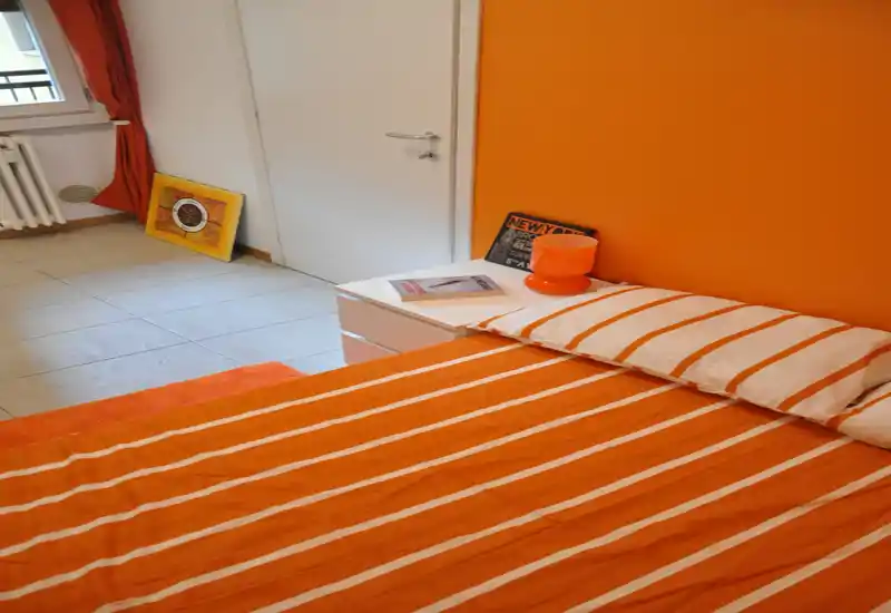 Camera da letto luminosa e moderna con copriletto a righe arancioni, cuscino abbinato e lampada sul comodino; pavimento piastrellato e arredi semplici per un ambiente caldo e contemporaneo.