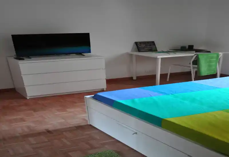 Camera da letto moderna e luminosa con un letto basso bianco con lenzuola colorate, un comò con TV e una scrivania semplice sullo sfondo. Linee pulite e pavimento in parquet danno una sensazione minimal e ariosa, perfetta per affitti brevi.