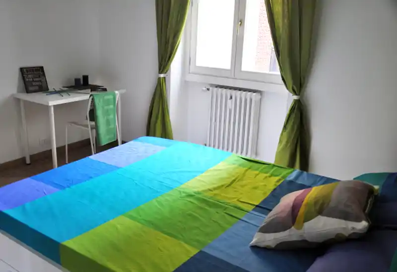 Camera da letto luminosa e ordinata con un ampio letto matrimoniale colorato che occupa la maggior parte dell'inquadratura, una scrivania vicino alla finestra e tende verdi che lasciano entrare luce naturale.