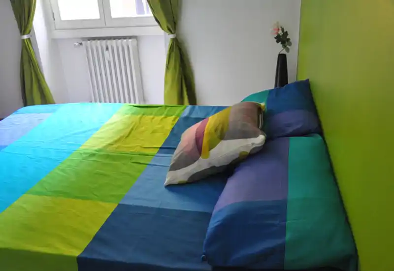 Camera da letto moderna e luminosa con un ampio letto dai colori vivaci; la luce naturale dalla finestra e un vaso decorativo semplice creano un’atmosfera fresca e accogliente.