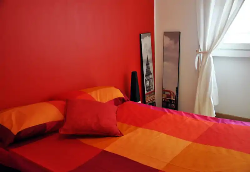 Camera da letto luminosa e accogliente con un grande letto con copriletto rosso-arancione, cuscino decorativo e luce naturale dalla finestra con tenda — ideale per annunci.