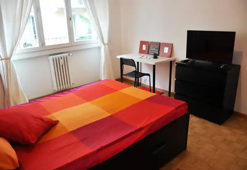 Camera da letto luminosa e ordinata con un letto matrimoniale con biancheria colorata, una piccola scrivania vicino alla finestra e una TV su un comò.