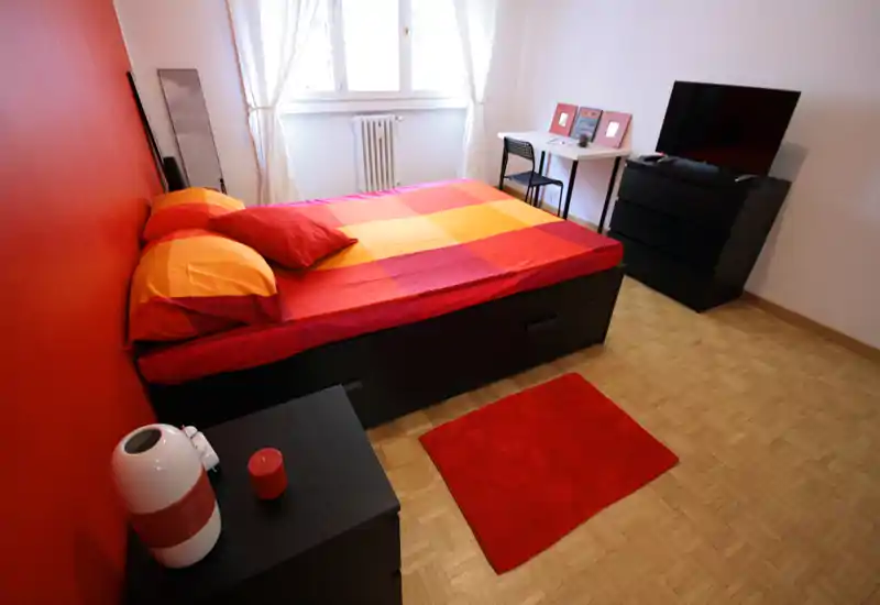 Camera da letto luminosa e ordinata con un grande letto con biancheria rossa e arancione, comodino, piccolo scrittoio e cassettiera con TV—ideale per un soggiorno accogliente in città.