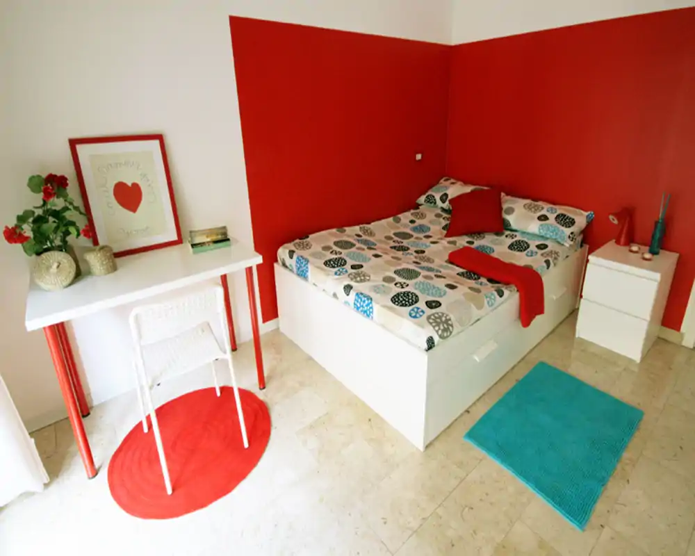 Camera da letto luminosa e ben arredata con letto matrimoniale a fantasia, pareti rosse a contrasto e tappeti coordinati per un aspetto moderno e accogliente.