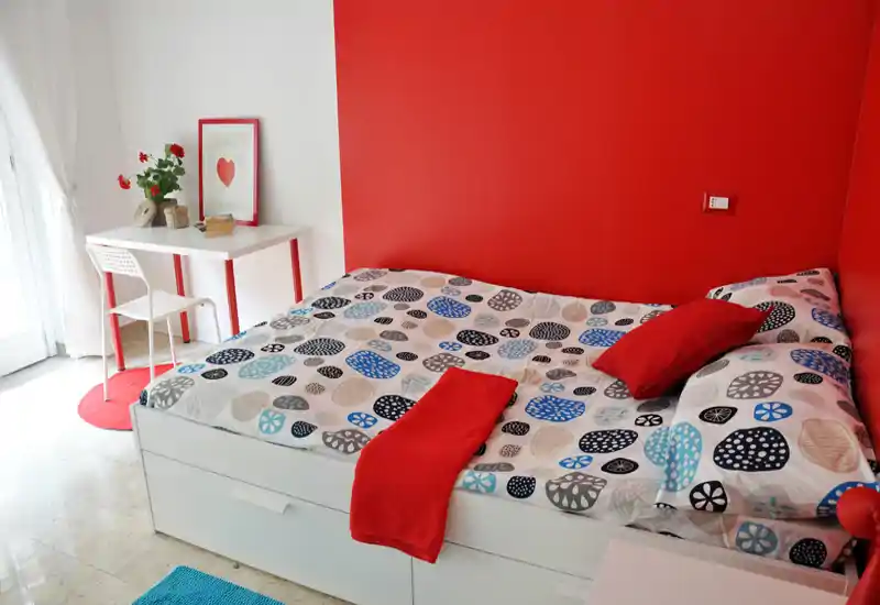 Camera da letto luminosa e moderna con letto matrimoniale, copriletto colorato e parete accentuata in rosso. Un piccolo angolo scrivania vicino alla finestra aggiunge funzionalità e luce naturale rende l’ambiente accogliente.