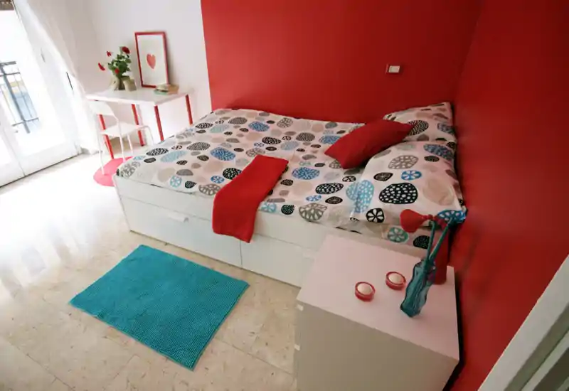 Camera da letto luminosa e ben arredata con letto matrimoniale, biancheria colorata e una piccola scrivania vicino al balcone che illumina naturalmente l'ambiente — ideale per un affitto accogliente.