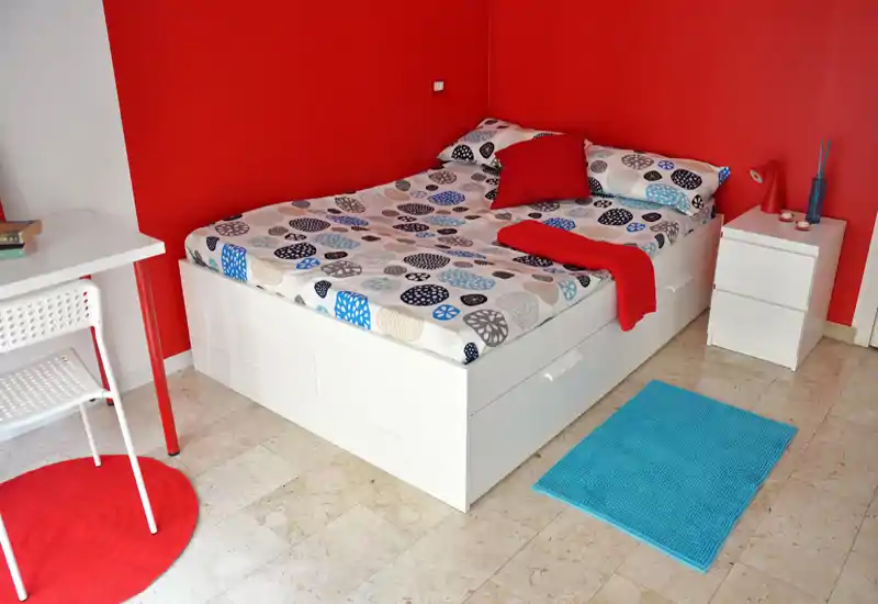 Camera da letto moderna e luminosa con letto matrimoniale, biancheria colorata e mobili bianchi minimal. Pareti rosse come accento, comodino e piccolo scrivania rendono l'ambiente accogliente e contemporaneo.