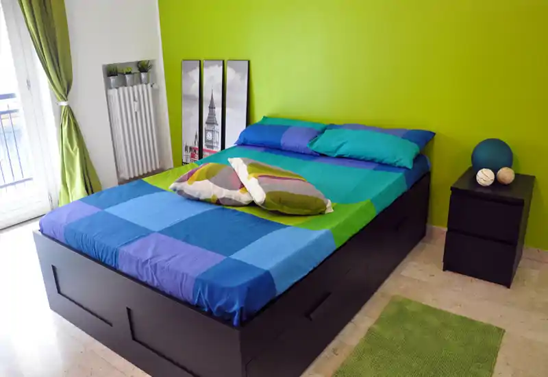 Camera da letto luminosa e colorata con un ampio letto matrimoniale, cuscini decorativi e comodino. La stanza ha pareti verde brillante e luce naturale proveniente dalla porta finestra.