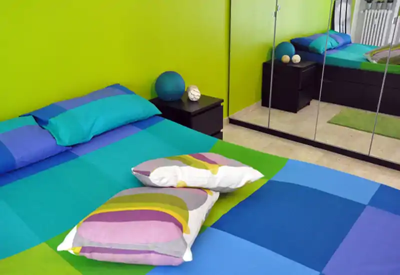 Camera da letto luminosa e colorata con un grande letto ben rifatto con biancheria blu e verde e cuscini decorativi; l'armadio specchiato riflette la stanza aumentandone la percezione di spazio.