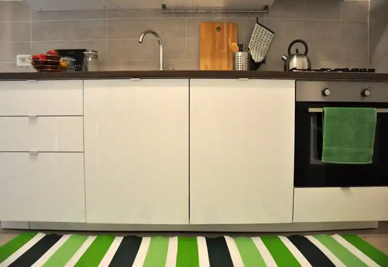 Cucina moderna compatta con mobili bianchi, lavello, forno incassato e dettagli verdi su asciugamano e tappeto — ordinata e luminosa.