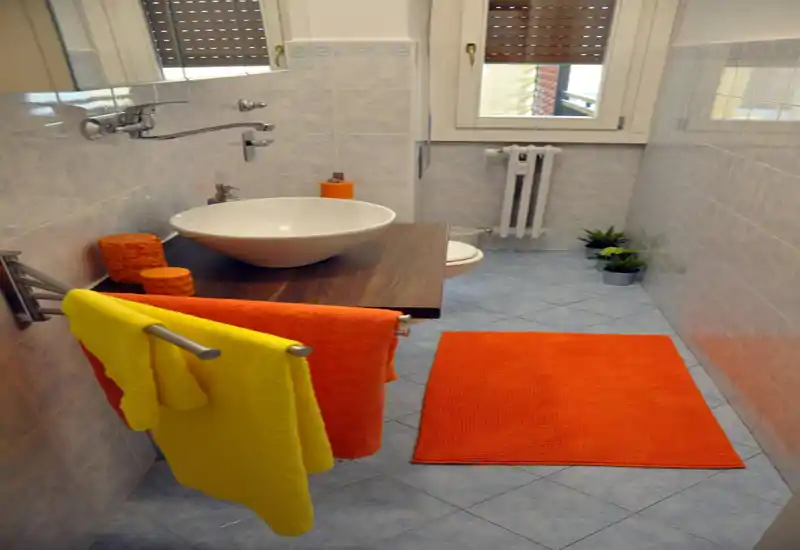 Bagno piastrellato di piccole dimensioni con lavabo a ciotola su mobiletto in legno, rubinetto a muro, porta asciugamani con asciugamani colorati e tappeto arancione; luce naturale dalla finestra e piante decorative vicino al WC.