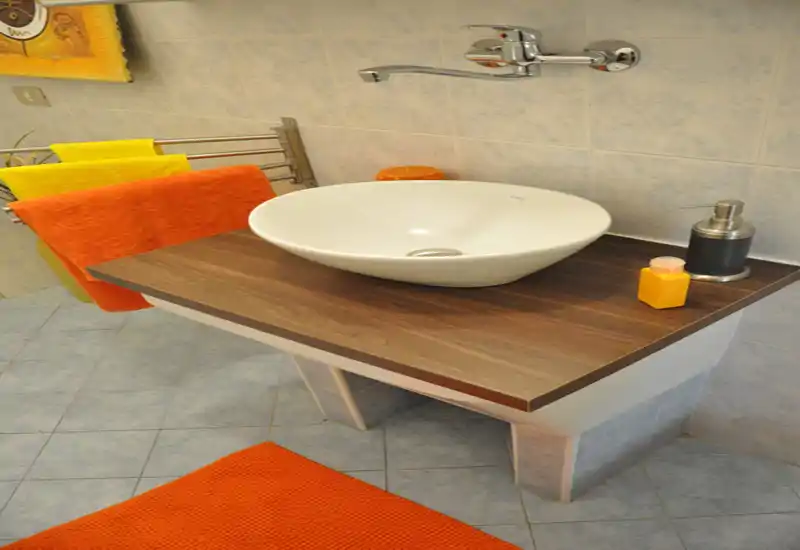 Lavabo moderno con bacinella su piano in legno, rubinetto a parete, dispenser per sapone e asciugamani colorati che aggiungono calore.