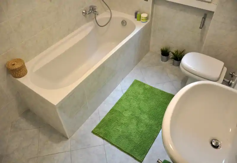 Bagno pulito e ben illuminato con vasca ad incasso, bidet/wc, lavabo e tappetino verde; piastrelle chiare che donano un aspetto fresco e neutro.