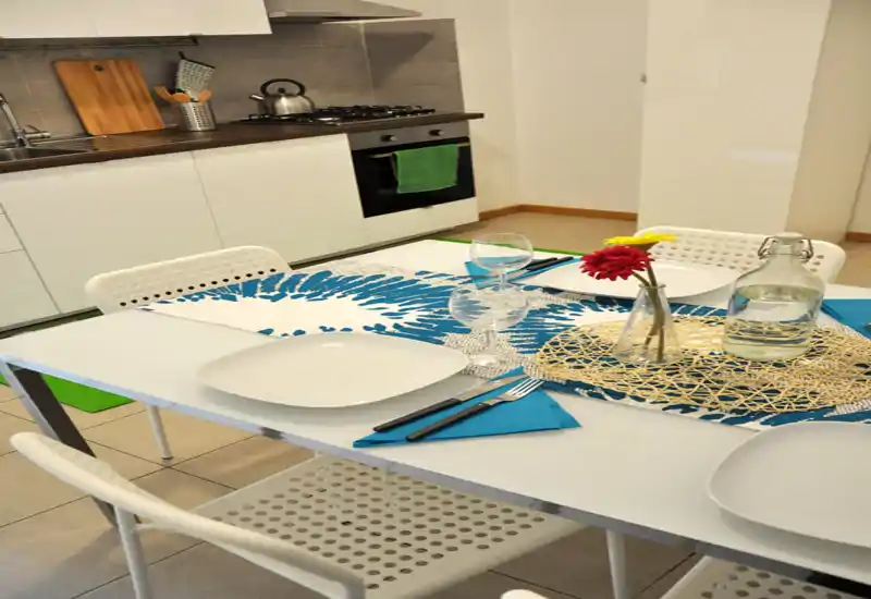 A neatly set dining table within an open kitchen, featuring modern white chairs, blue placemats, glassware and a decorative centerpiece — ideal for a cozy meal.
