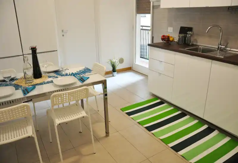 Cucina moderna e luminosa con un piccolo tavolo da pranzo per quattro persone, mobili bianchi, lavello in acciaio e un tappeto a righe verde. Spazio ordinato e ben illuminato, ideale per un annuncio.